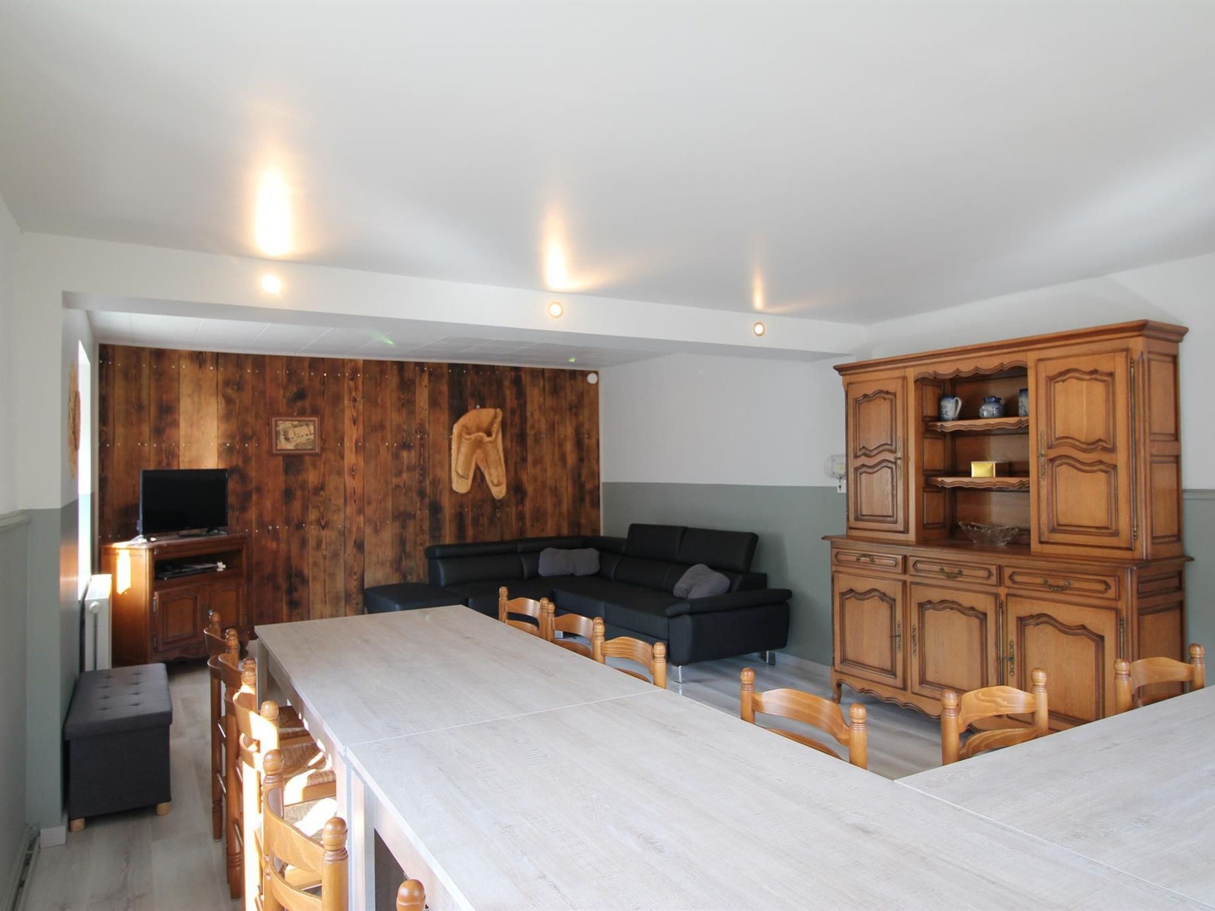 A dining area with a long table and chairs, a wooden cabinet with shelves, a television on a stand, and a wall with wooden panels and a hanging pair of boots.