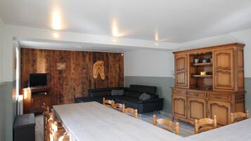 A dining area with a long table and chairs, a wooden cabinet with shelves, a television on a stand, and a wall with wooden panels and a hanging pair of boots.