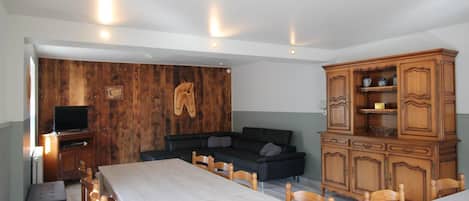 Un comedor con una mesa larga y sillas, un mueble de madera con estantes, un televisor sobre un soporte, y una pared con paneles de madera y un par de botas colgadas.