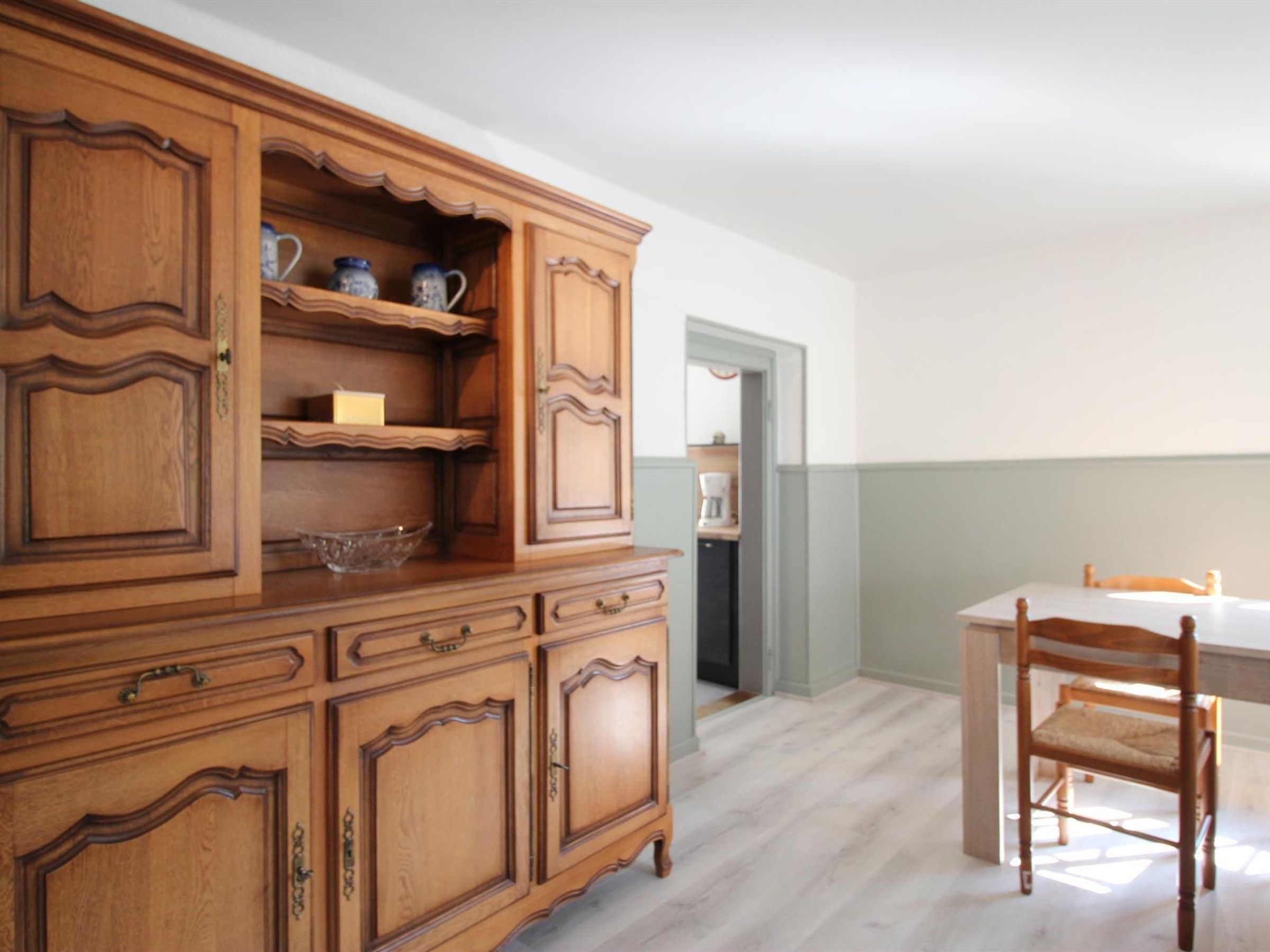 Ein Holzschrank mit vielen Schubladen und Regalen, ein Esstisch mit Stühlen und im Hintergrund ein Küchenbereich.