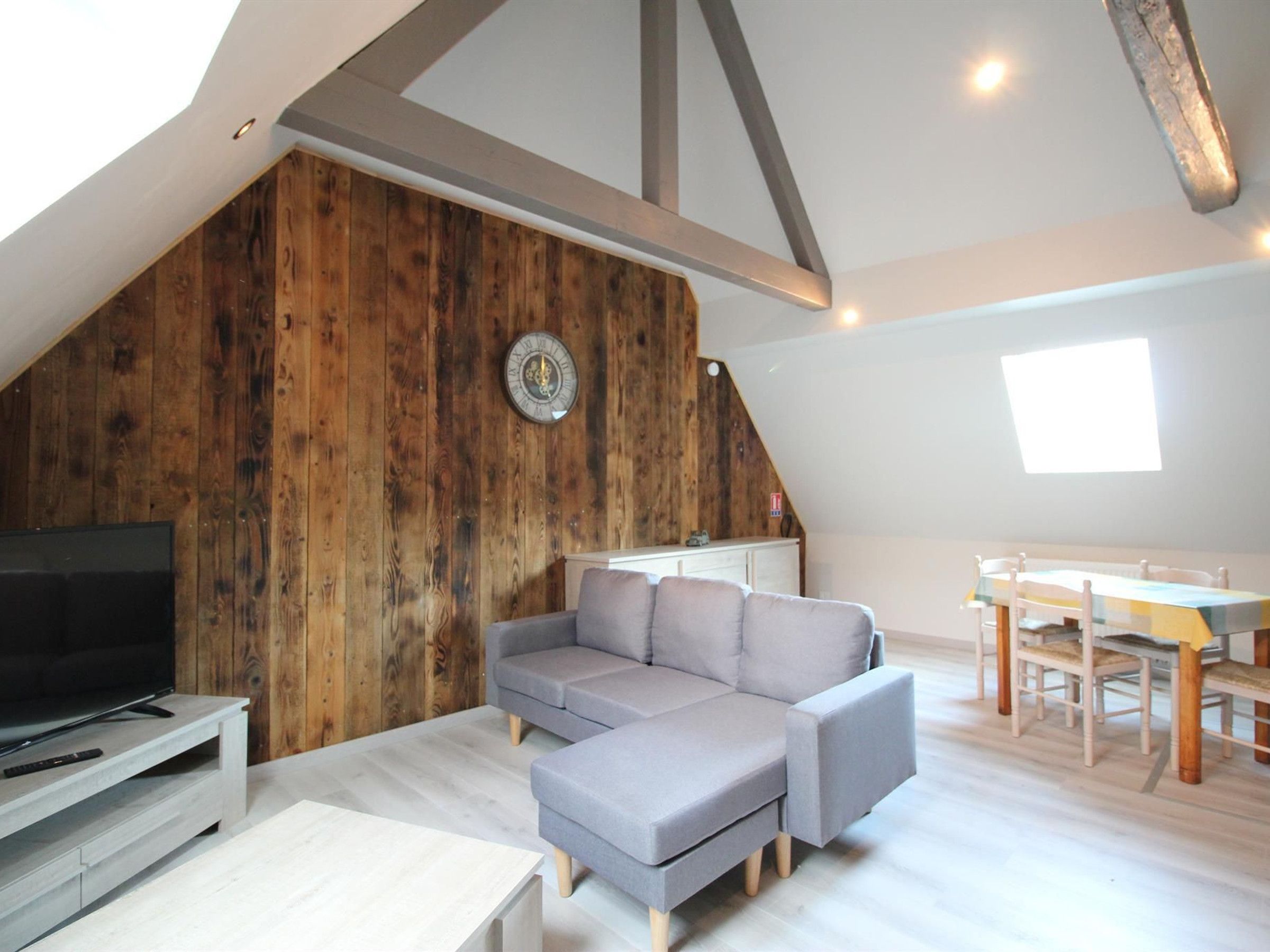 Ein Loft-Wohnzimmer mit einer Holzvertäfelung, einem grauen Sofa, einem Fernseher auf einem tiefen Schrank und einem Esstisch mit Stühlen.