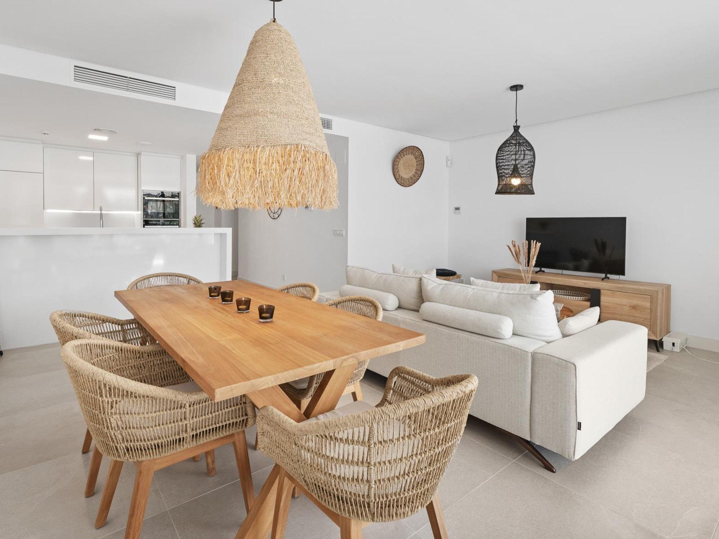 A modern living space with a wooden dining table, wicker chairs, a sofa, and a television.
