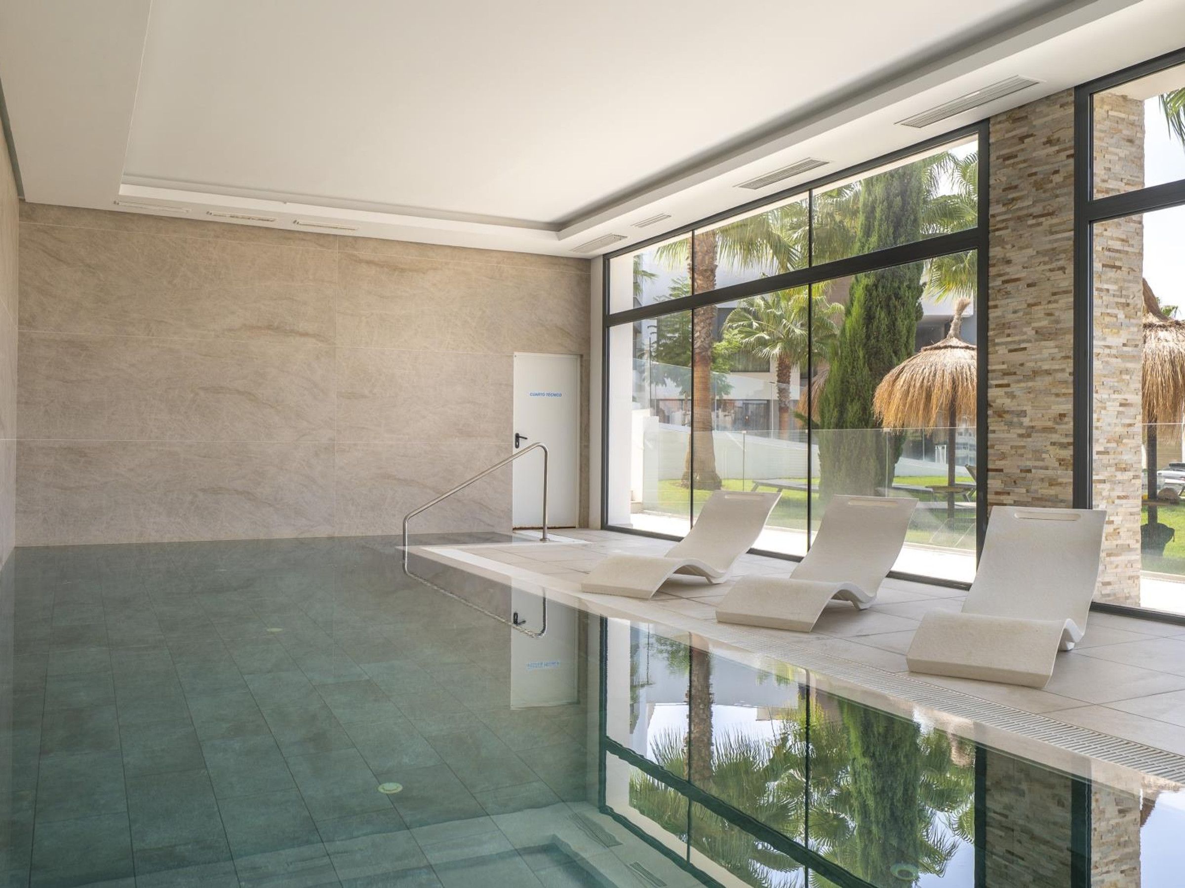 Un espace piscine intérieur moderne, doté de murs en verre transparent, d’un sol carrelé et de chaises longues.