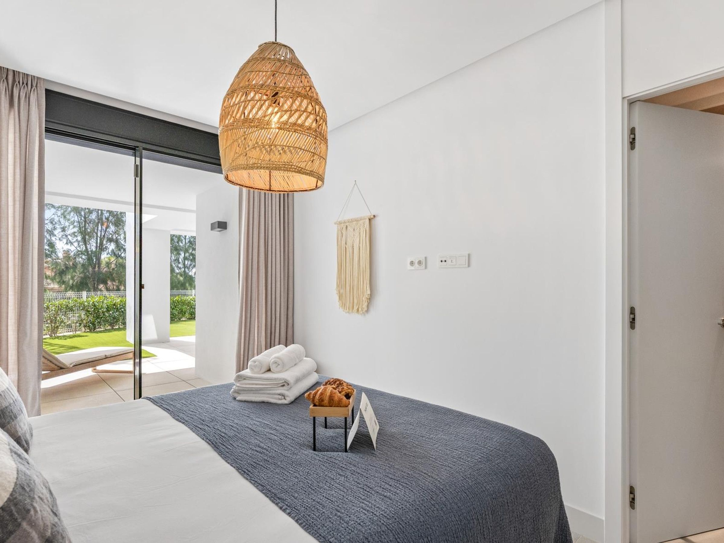 A modern bedroom with a bed, a hanging light fixture, a small table with a croissant, and a view of a garden through a sliding door.