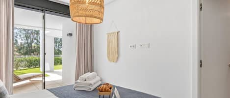 A modern bedroom with a bed, a hanging light fixture, a small table with a croissant, and a view of a garden through a sliding door.