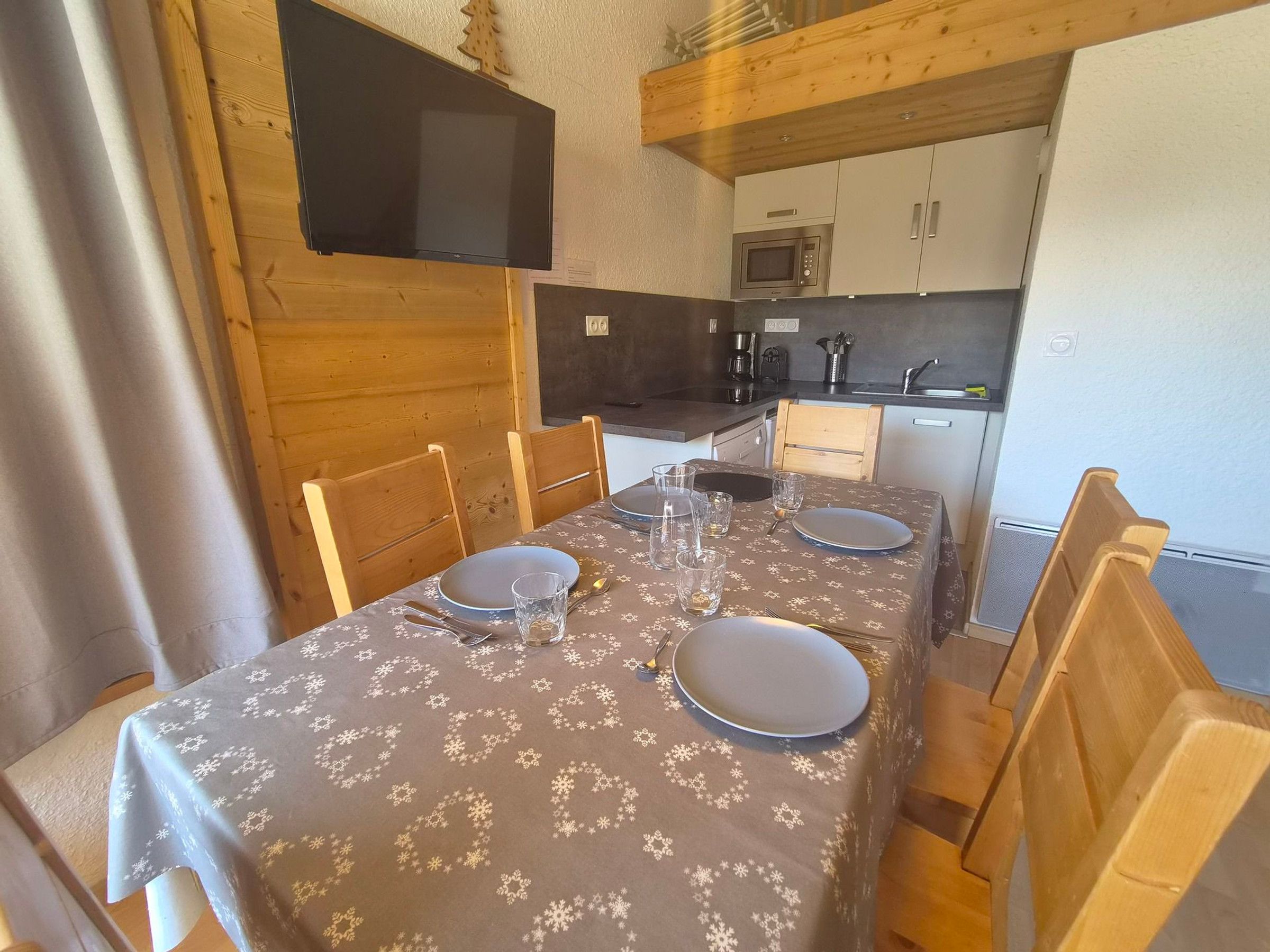 Ein Essbereich mit Tisch für vier Personen, Holzsesseln, einem an der Wand befestigten Fernseher und einer Küchenzeile mit Schränken und Geräten.