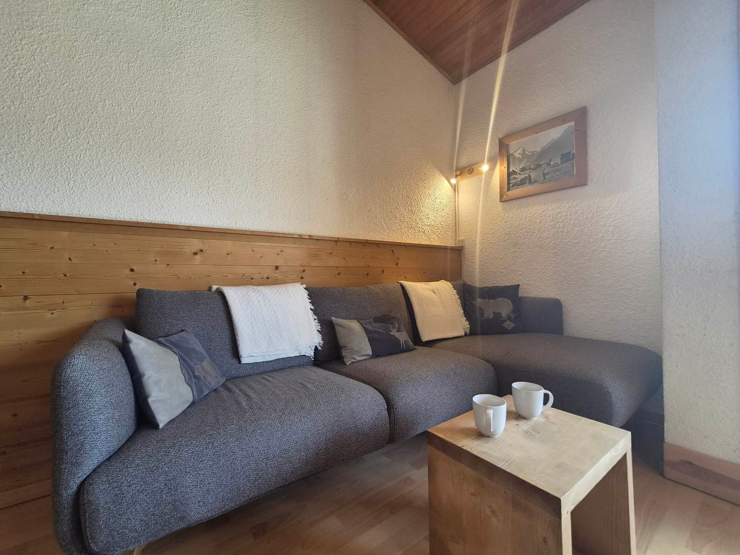 Une pièce chaleureuse avec une tête de lit en bois, un canapé gris agrémenté de coussins et une petite table en bois sur laquelle sont posées deux tasses blanches.