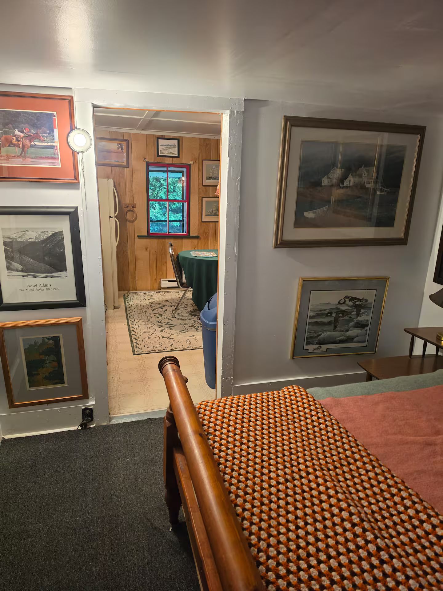 A room with a bed, framed pictures on the walls, a window, and a door leading to another room.