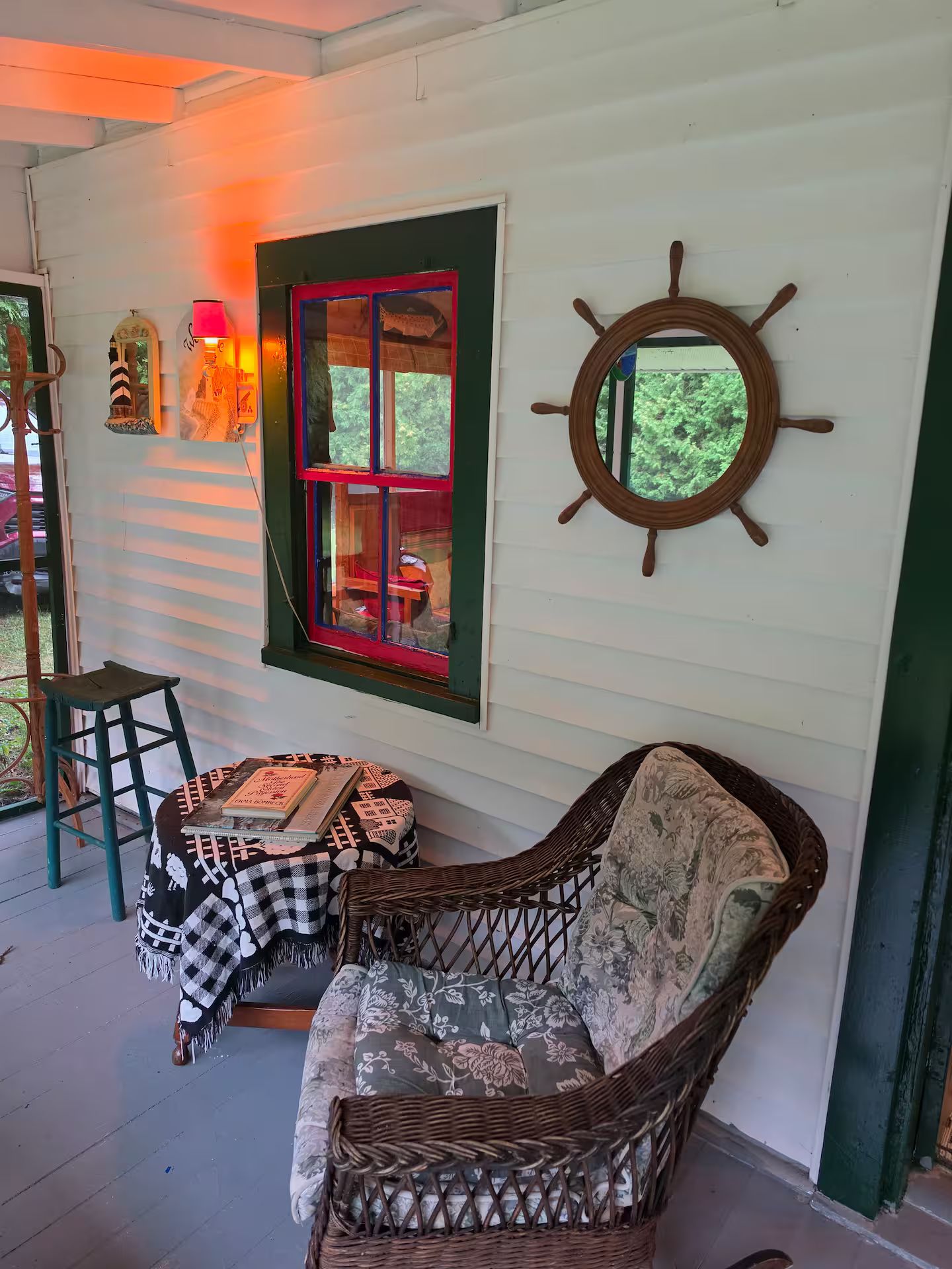 A cozy room with a wicker chair, a table with books, a window with red trim, and a ship's wheel decoration.