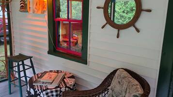 A cozy room with a wicker chair, a table with books, a window with red trim, and a ship's wheel decoration.