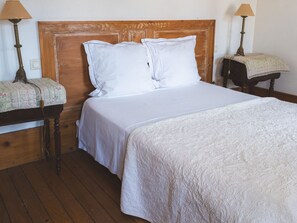 A bedroom with a wooden headboard, two white pillows, a white bedspread, and a bedside table with a lamp and a floral-patterned tablecloth. - Charming quiet cottage - 2 guests (Choloy-Ménillot)