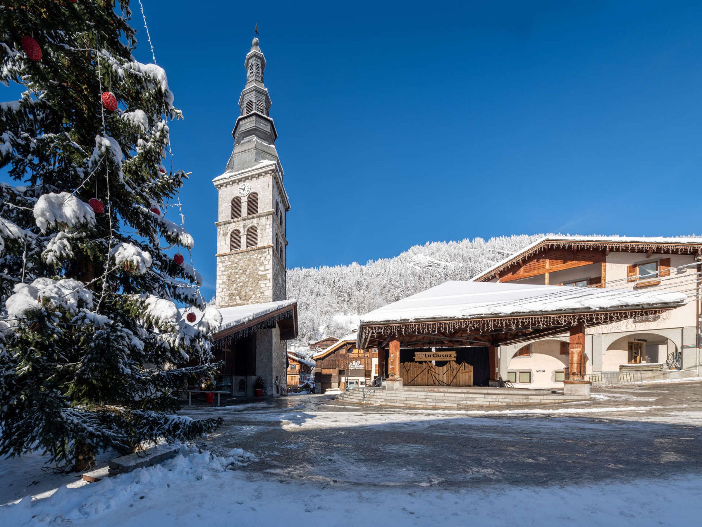 A snow-covered building with a clock tower, a sign reading 'La Clusaz,' and a snow-laden tree.