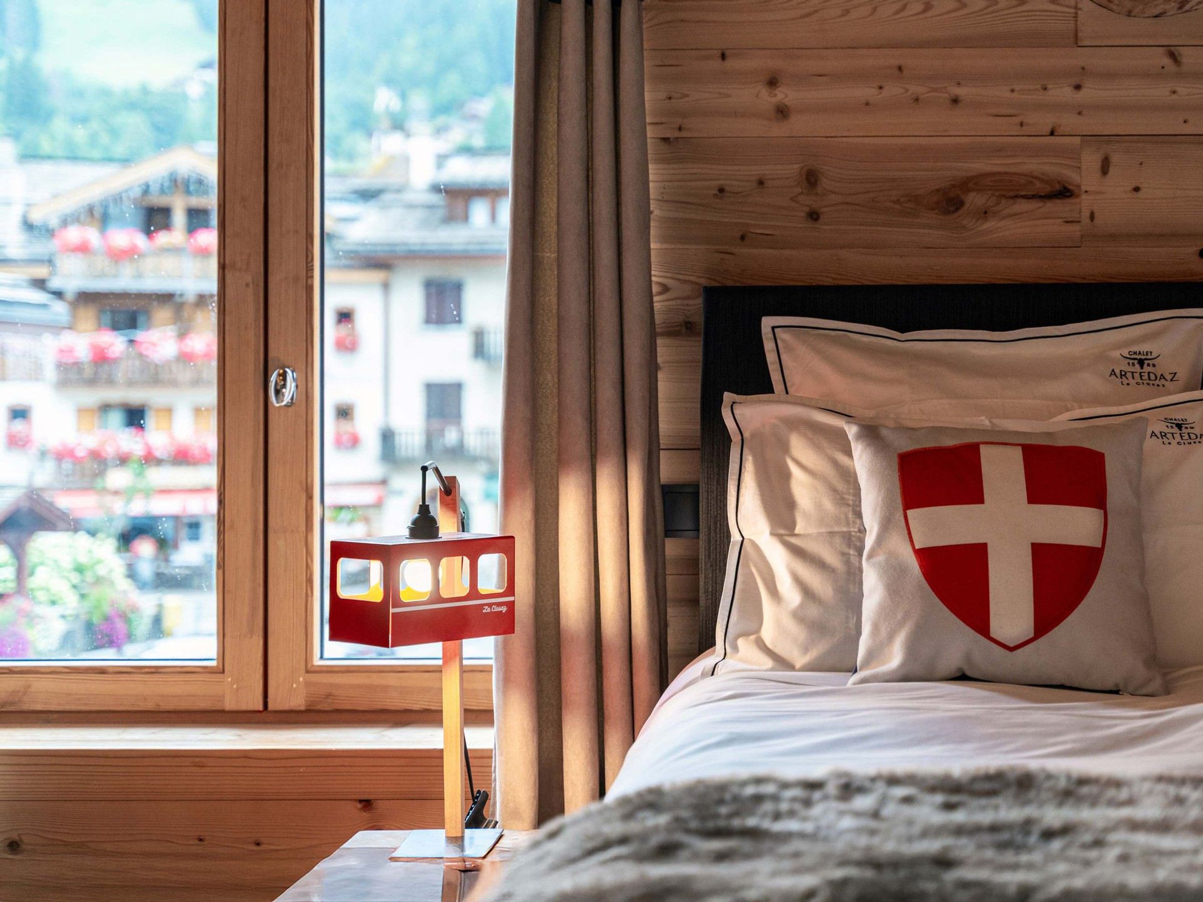 A hotel room with a wooden headboard, a window with a view of buildings, and a red and white pillow with a cross emblem.