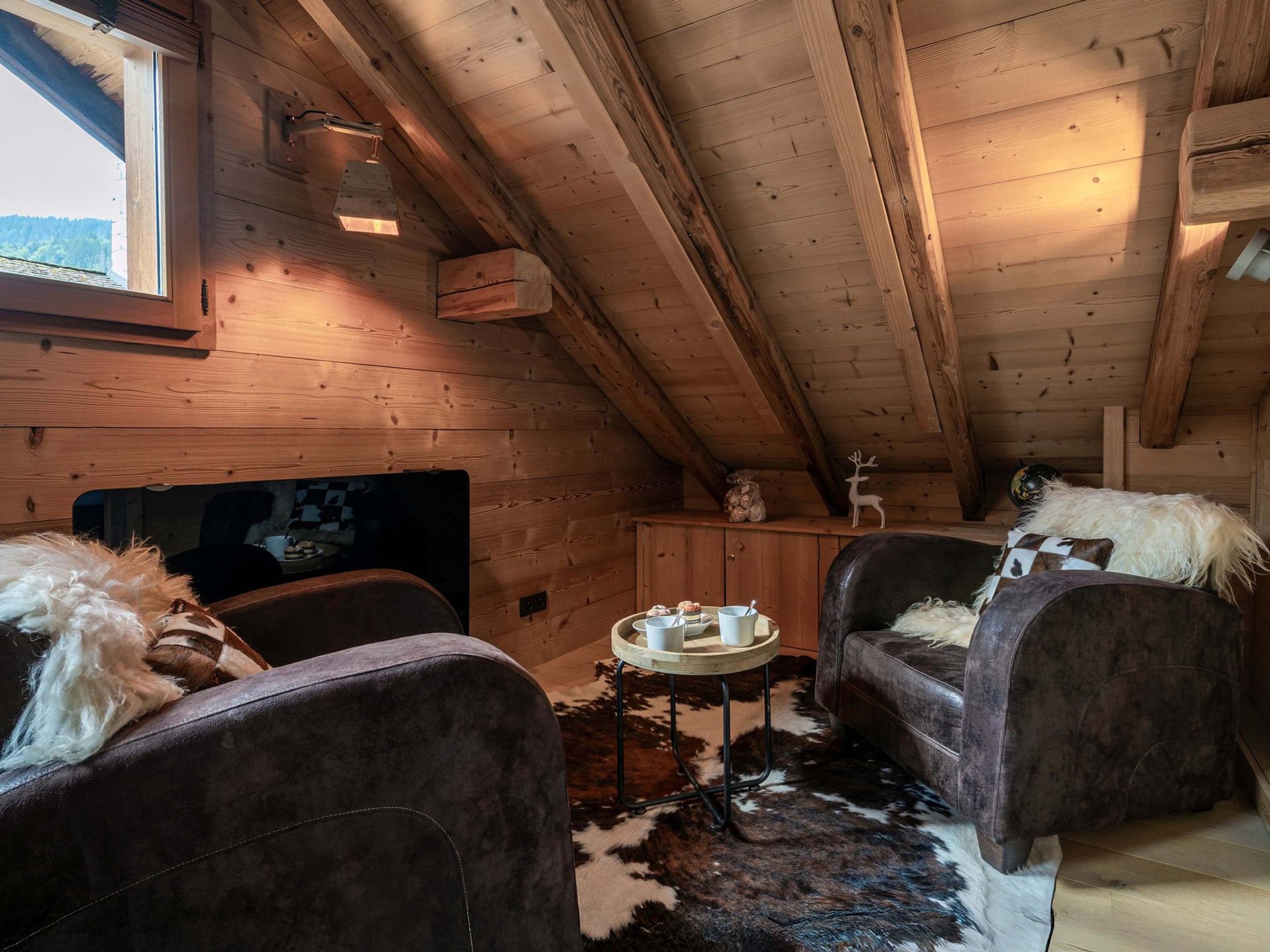 Interior acogedor de una cabaña con paredes de madera, techo inclinado y decoración rústica que incluye un sofá cubierto de piel, una pequeña mesa redonda con tazas y una figurina de ciervo.