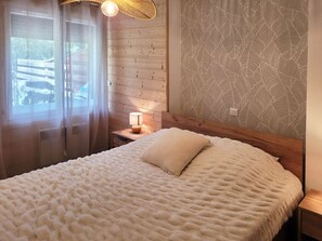 Ein Schlafzimmer mit einem hölzernen Kopfteil, einem Bett mit weißer, strukturierter Decke, einer Nachttischlampe und einem Fenster mit Blick ins Freie.