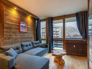 A modern living room with a grey sectional sofa, a wooden coffee table, and a large window with a view of buildings and a forested area. - Elegant apartment, pets welcome - 4 guests (Val-d'Isère)