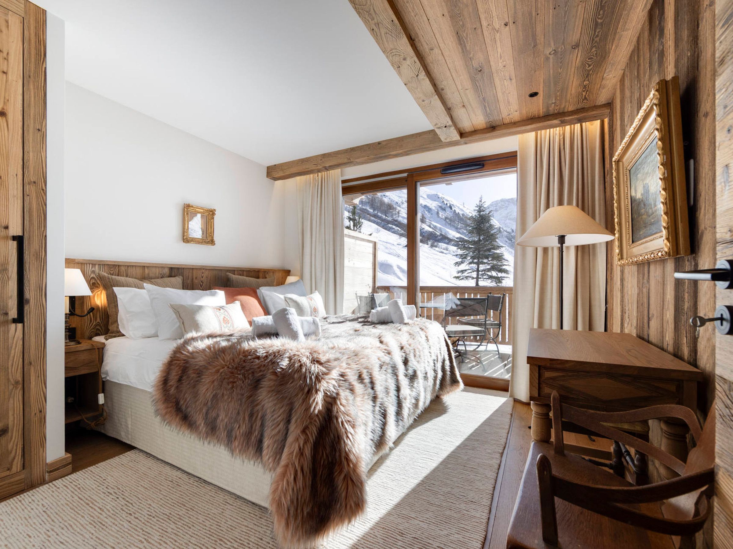 A bedroom with a large bed, wooden headboard, a view of mountains, a wooden desk, and a chair.