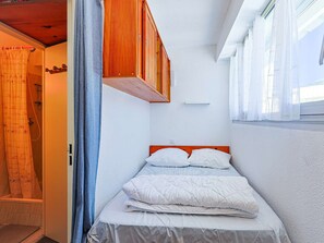 Ein kleines Schlafzimmer mit einem Einzelbett, einem Holz-Kopfstück, einem Fenster mit durchsichtigen Vorhängen und einer Duschkabine mit Vorhang.