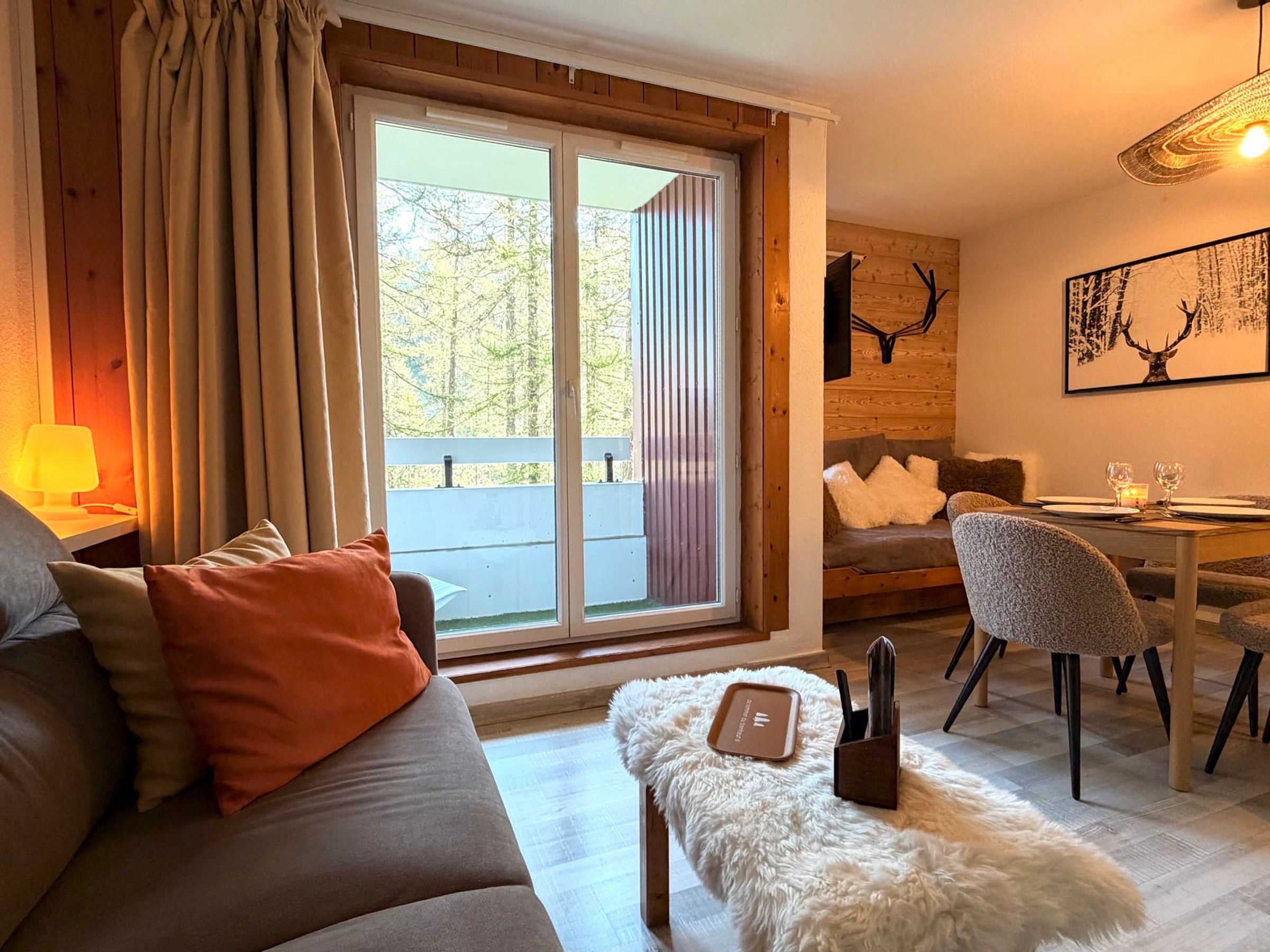 A cozy living room with a sofa, a dining table with chairs, a fireplace, and a large window with curtains.