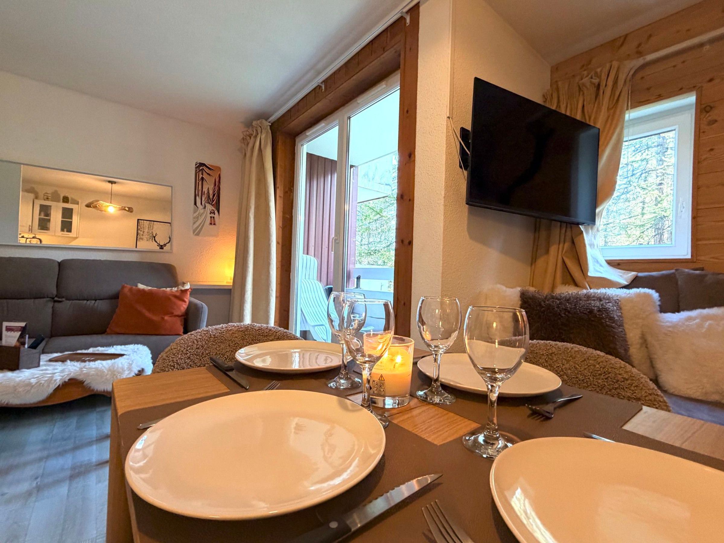 A dining area with a table set for two, a television mounted on the wall, and a sofa in the background.