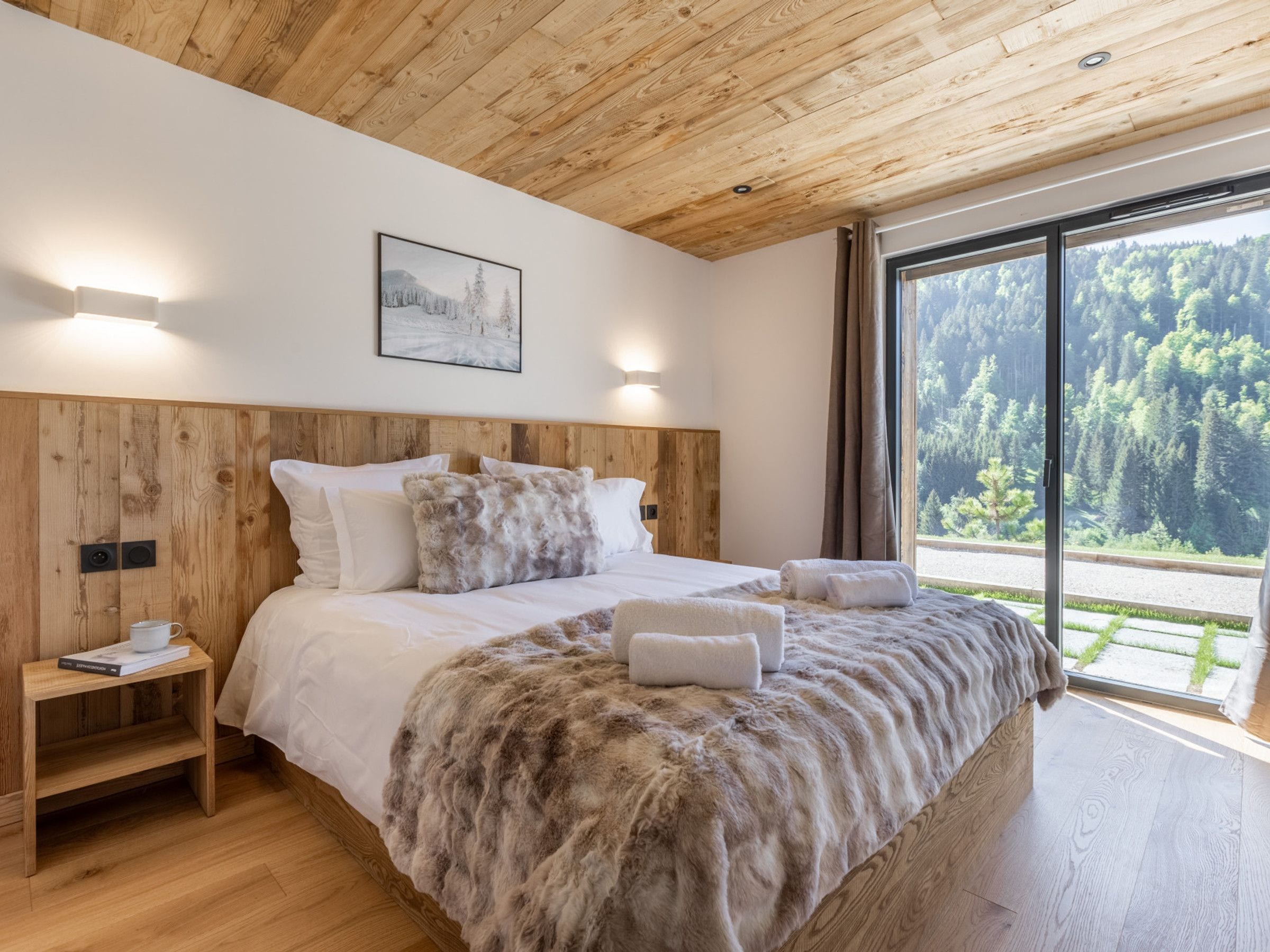 A bedroom with a wooden headboard, a large bed with white bedding, a fluffy throw blanket, a bedside table with a book, and a window with a forest view.