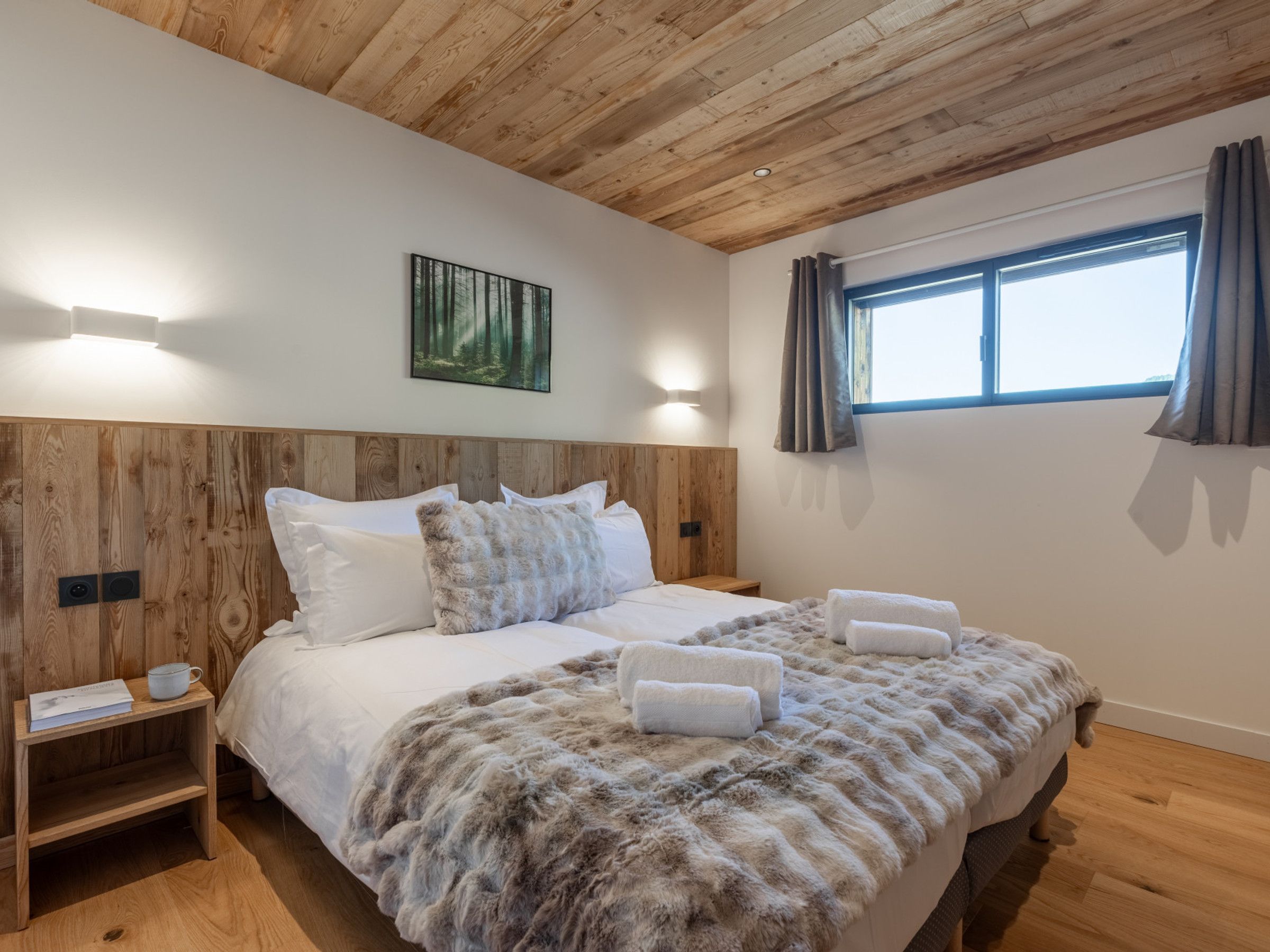 A bedroom with a wooden headboard, a large bed with white bedding, a fluffy blanket, two bedside tables with a cup and book, and a painting on the wall.
