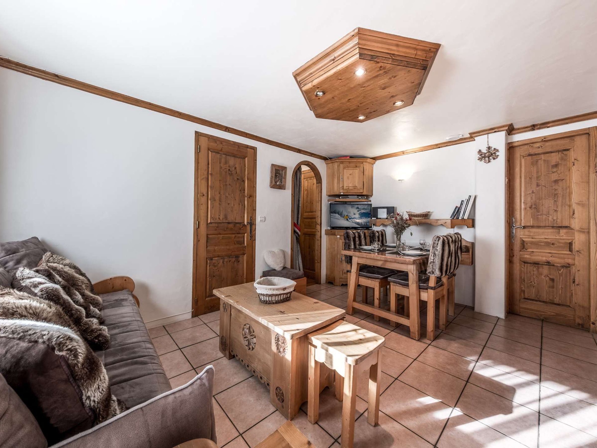 A cozy living room with a sofa and a wooden coffee table, a kitchen area with wooden cabinets and a dining table, and a ceiling with a wooden beam design.