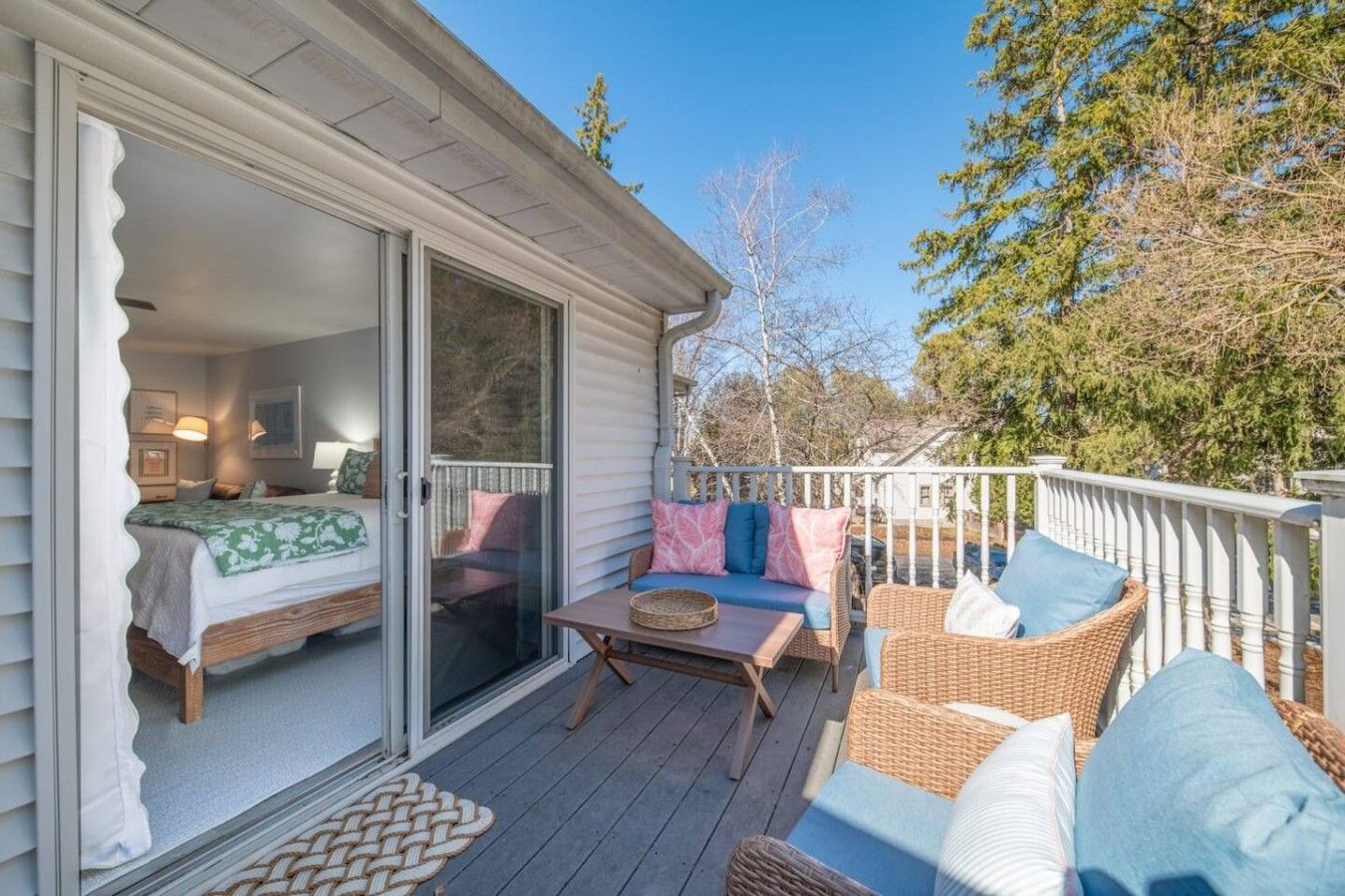 Une terrasse couverte avec des meubles d’extérieur, une porte-fenêtre coulissante donnant sur une chambre, et une vue sur les arbres et le ciel.