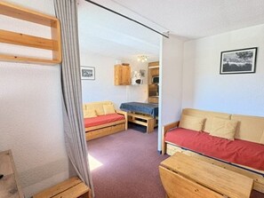 Ein kleines Zimmer mit zwei Betten, einer hölzernen Bank, einem kleinen Tisch und einer Küchenzeile im Hintergrund.