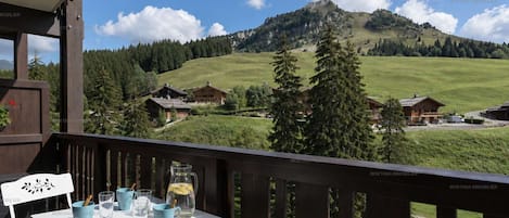 A balcony with a table set for two, overlooking a mountainous landscape with houses and trees.