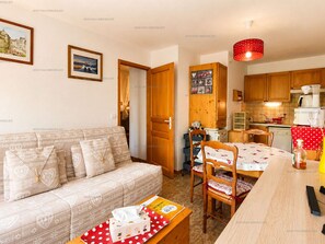 A cozy living room with a sofa, a dining table with chairs, and a kitchen area with wooden cabinets and a red hanging light. - Cozy apartment with mountain view - 4 guests (Le Grand-Bornand)