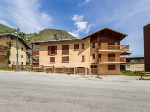 A multi-story building with balconies, a paved ground floor, and a mountainous backdrop. - Cozy apartment - 4 guests (Valloire)
