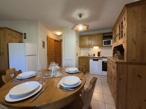 A dining area with a wooden table set for four, a white refrigerator, wooden cabinets, and a microwave in the kitchen area. - Practical apartment, private parking - 6 guests (Praz-sur-Arly)