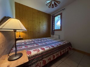 A bedroom with a wooden headboard, a bed with patterned bedding, a bedside lamp, a window with curtains, and a radiator. - Practical apartment, private parking - 6 guests (Praz-sur-Arly)