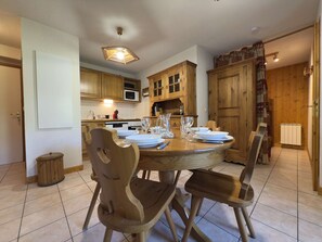 A dining area with a round wooden table set for four, surrounded by wooden chairs. There is a kitchen in the background with wooden cabinets and a microwave. - Practical apartment, private parking - 6 guests (Praz-sur-Arly)