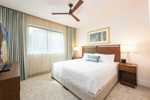 Une chambre d’hôtel comprenant un lit, un bureau, une chaise, un ventilateur de plafond, une fenêtre avec des rideaux et un tableau encadré au mur.