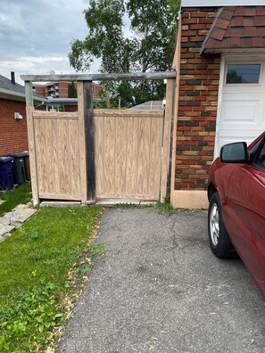 A residential driveway with a wooden fence, a red car parked on the right, and a brick house with a garage door. - A large, very comfortable private room just 10 minutes from the subway. (Laval)