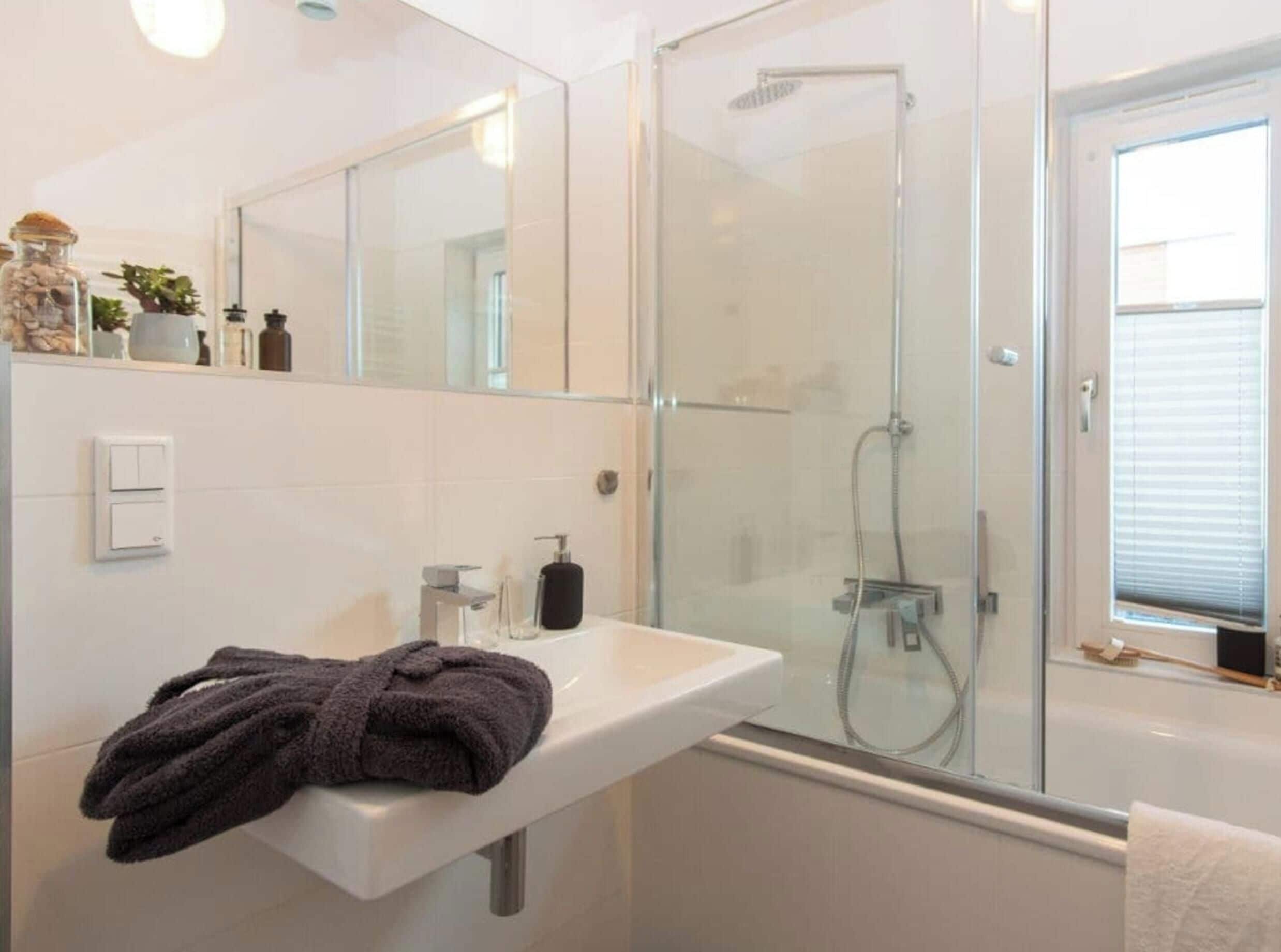 Ein modernes Badezimmer mit einer in Glas abgeschirmten Dusche, einem weißen Waschbecken mit Handtuch und einem Spiegel darüber.