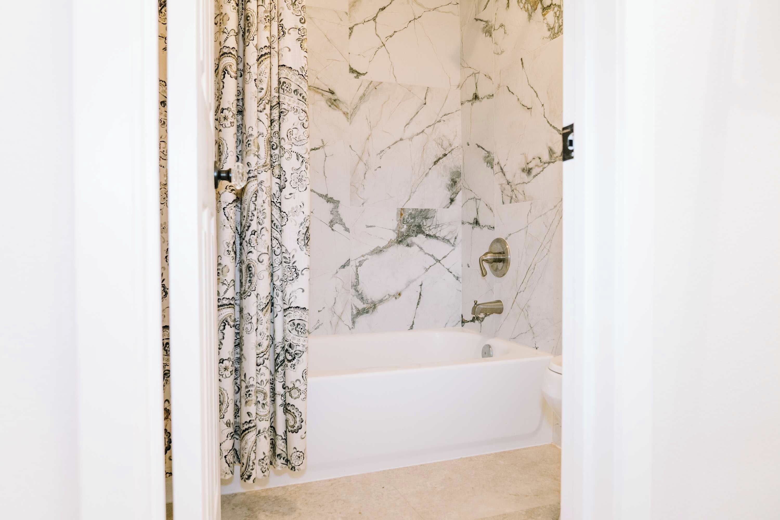 A bathroom with a marble wall, a white bathtub, and a patterned shower curtain.
