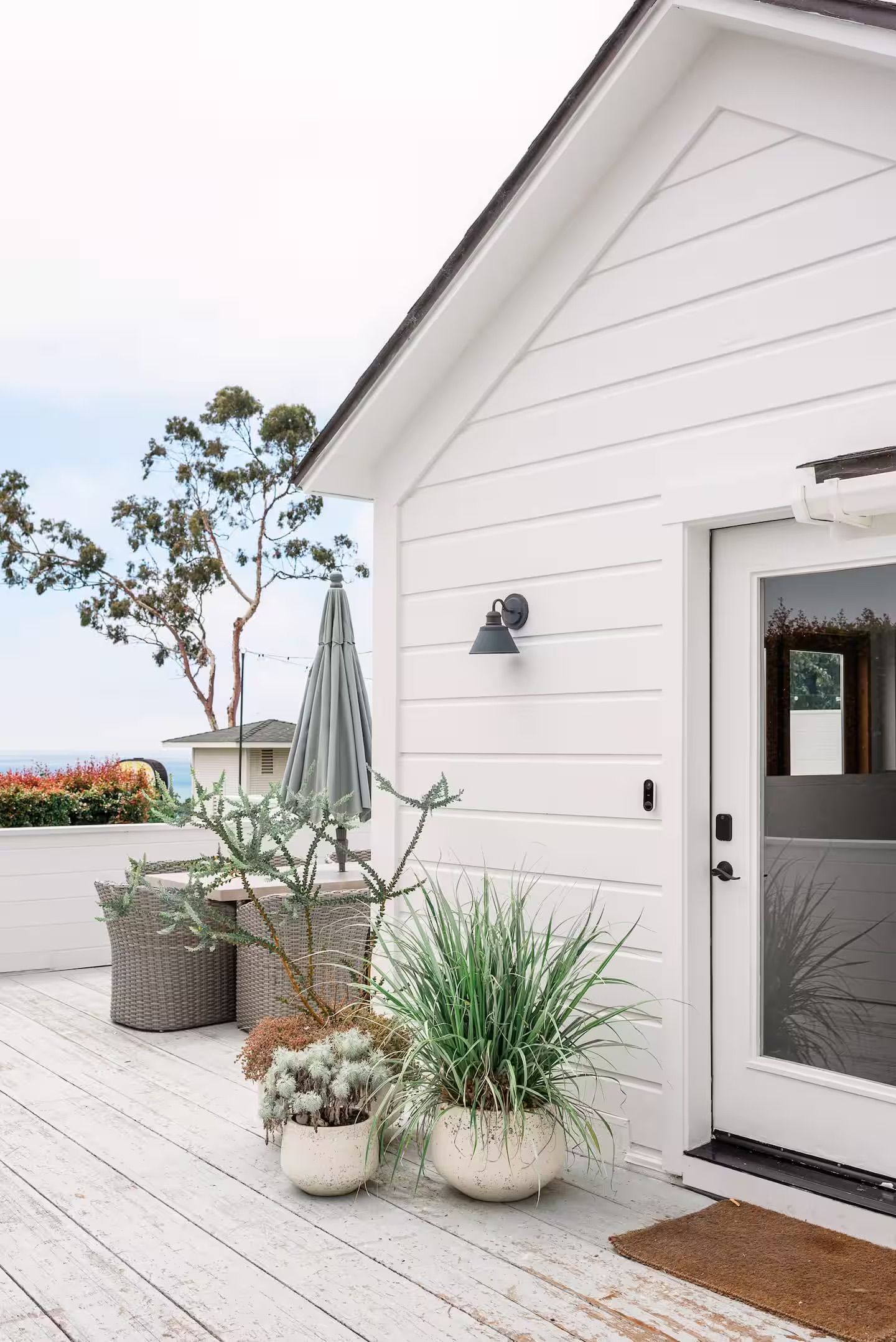 A white building with a door, a small porch with potted plants, and a grey umbrella.