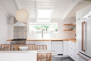 A modern kitchen with white cabinetry, a large window, and a dining area with wicker chairs. - Summerland Panoramic Ocean & Island Views – Walk to Beach & Town - 4Bed 4.5 Bath (Summerland)