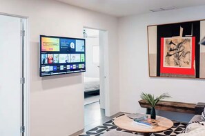 Ein modernes Wohnzimmer mit einem an der Wand befestigten Flachbildfernseher, der verschiedene Apps und Kanäle anzeigt. Es gibt einen runden Couchtisch mit Büchern und einer kleinen Pflanze, sowie ein gerahmtes abstraktes Gemälde an der Wand.