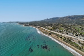 An aerial view of a coastal area with a beach, a highway, and a mountain range in the background. - Summerland Panoramic Ocean & Island Views – Walk to Beach & Town - 4Bed 4.5 Bath (Summerland)
