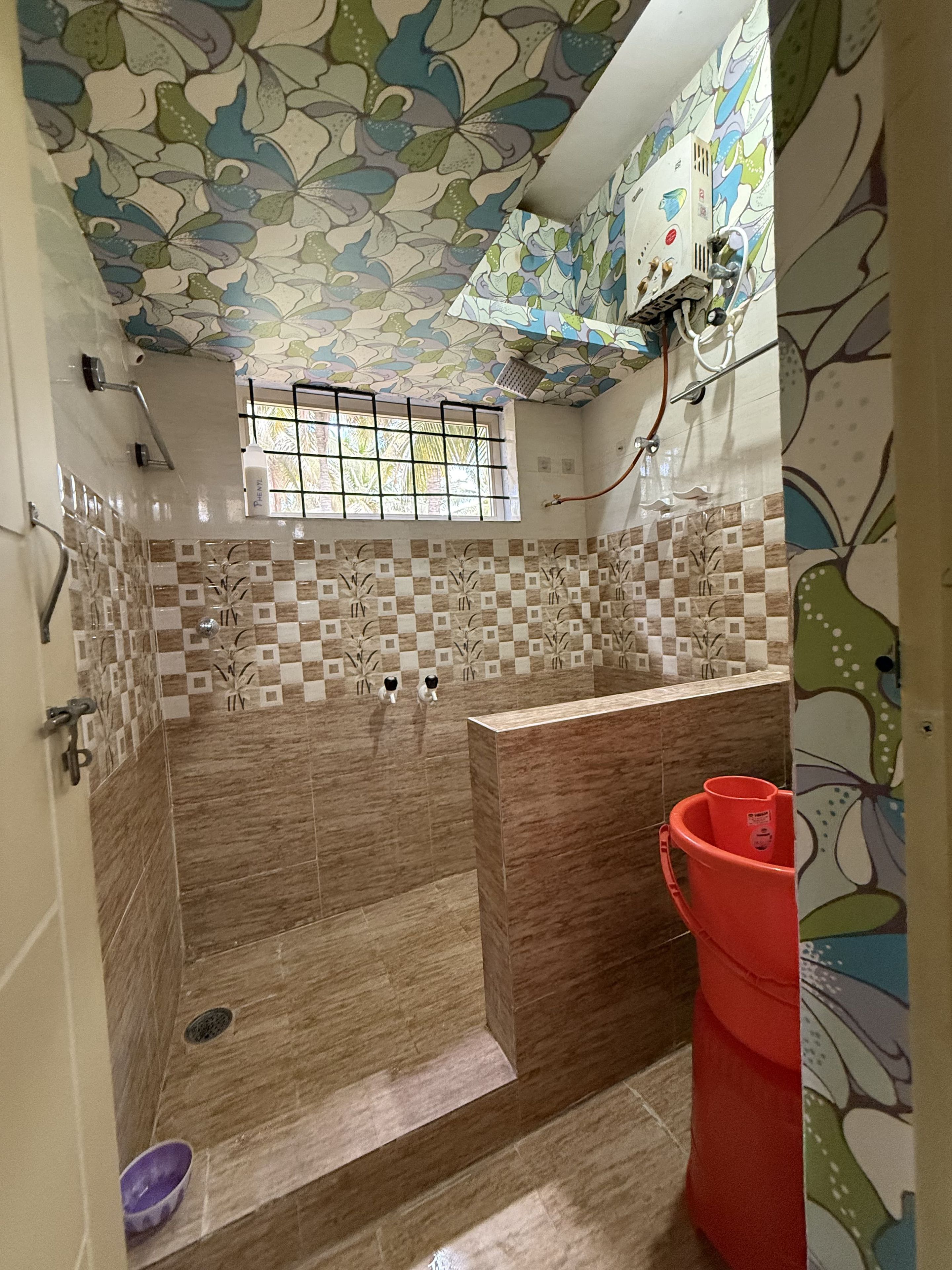 Un baño con ducha, paredes de azulejos, una ventana y un cubo rojo.