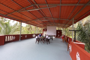 An outdoor covered area with tables and chairs, surrounded by red walls and palm trees. - Coconut Sooru Homestay Channapatna (Kurnagere)