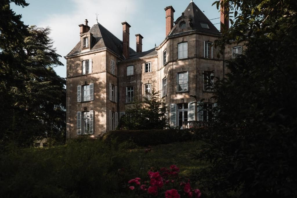Un grand bâtiment historique, doté de nombreuses cheminées et fenêtres, entouré d’une végétation luxuriante et d’un jardin où fleurissent des plantes aux couleurs roses.
