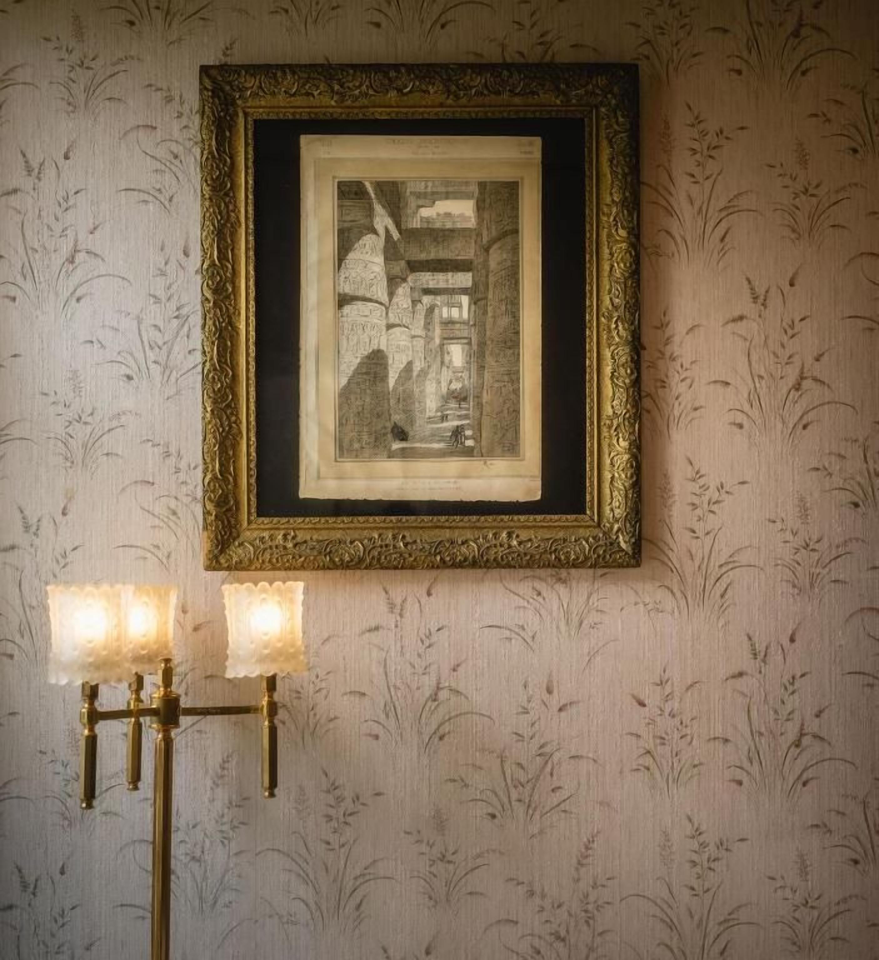 A framed black and white photograph on a wall with a patterned wallpaper background. Below the photo, there is a wall-mounted brass candelabra with three lit candles.