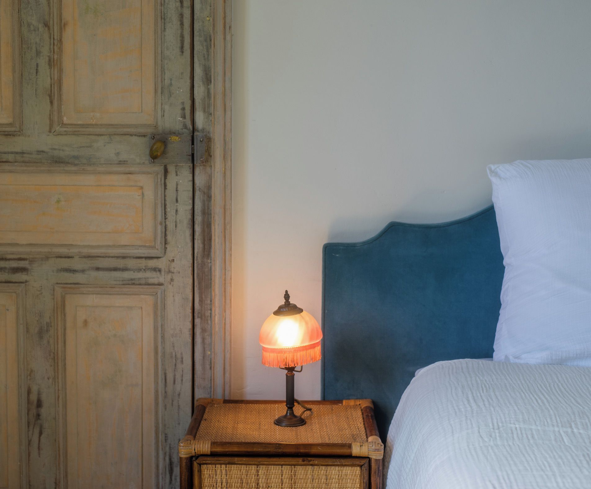 Ein Nachttisch aus Holz mit einer Lampe, ein Bett mit blauer Kopfteil und eine Holztür.