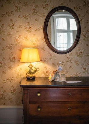 A wooden dresser with a mirror above it, a lamp on the dresser, and a floral wallpaper background. - Château de Sainte-Sévère (Sainte-Sévère-sur-Indre)