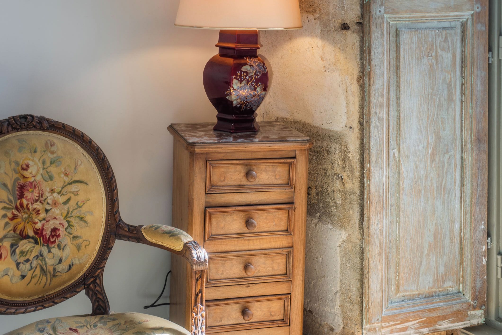 A wooden dresser with a floral-patterned lamp on top, a vintage chair with a floral design, and a wooden door.
